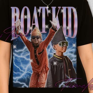 May include: Black t-shirt featuring a graphic of two figures in orange and black outfits, wearing sunglasses and hats, with the text "BOAT KID" in a purple and white font. The background includes a lightning effect.