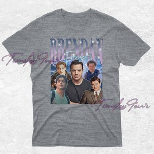 May include: Gray t-shirt featuring a collage of images of actor Brendan Fraser. The design includes the name "Brendan Fraser" in a stylized font with a light blue and purple gradient. The shirt is a classic crew neck.
