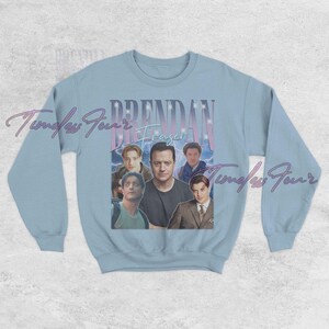 May include: Light blue sweatshirt featuring a collage of images of Brendan Fraser. The name "Brendan Fraser" is printed in a retro font above the images. The sweatshirt has long sleeves and a crew neck.