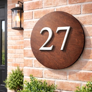 Puede incluir: Un letrero redondo de madera con el número "27" en blanco. El letrero está montado en una pared de ladrillo. El letrero es de color marrón oscuro y los números están en relieve. Una linterna negra es visible.