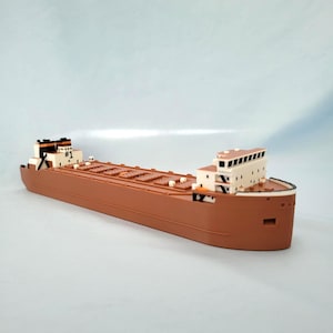May include: A brown model cargo ship featuring a cream-colored superstructure at the bow and stern. The stern cabin displays the text #1 in black. The deck is segmented with rectangular hatch covers, and the hull has a smooth, curved design.