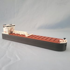 May include: A white and black model of a large freighter ship with a brown cargo hold. The ship has a raised bridge and a small white cabin on the bow.