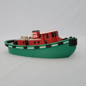 May include: A toy tugboat in green, red, and white. The boat has a green hull, a red cabin with black accents, and a white roof. The smokestack is red with a white 'G' on it. The boat is a miniature model.
