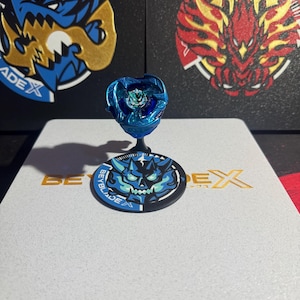 Może przedstawiać: Niebieska i srebrna zabawka Beyblade z czarno-białym logo Beyblade na białym tle. Logo zawiera napis "Beyblade".