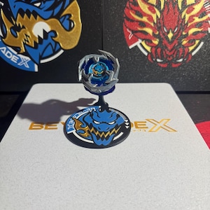 Op de afbeelding: Een blauw en zilveren Beyblade-speelgoed met een blauw en geel drakenontwerp op de basis. De Beyblade staat op een witte displaystandaard met de woorden "Beyblade X" in goud.