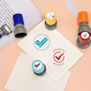 May include: A selection of self-inking stamps in various colours, including blue, orange, yellow, and red. The stamps feature text such as "Checked by Mrs. Miller", "From Mrs. Emma", and "Checked by Mrs. Chat".