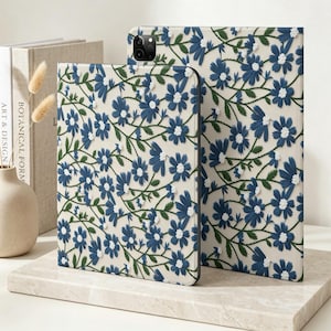 Peut inclure: Deux étuis pour tablette avec un fond crème et un motif répétitif de fleurs bleu foncé ressemblant à des marguerites, dotées de centres blancs et de fines tiges vertes. Les étuis ont un aspect texturé et une découpe pour l'appareil photo.