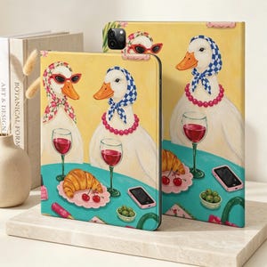 May include: Two tablet cases featuring a yellow background with an illustration of three white ducks wearing headscarves and necklaces. The ducks are seated at a teal table with glasses of red wine, croissants, cherries, olives, and smartphones.