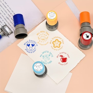 Pode incluir: Uma coleção de carimbos coloridos para professores com vários desenhos e frases, incluindo um sol sorridente, uma estrela e uma maçã. Os carimbos são azuis, amarelos e vermelhos, com cabos cinzentos. Frases como "Proud of you!" e "Great Job!"
