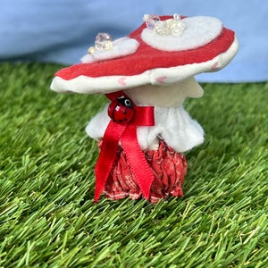 May include: A charming mushroom ornament featuring a red and white cap decorated with clear beads. The mushroom has a white stem, a red skirt, and a red bow with a ladybird detail. It is set against a backdrop of green grass and a blue sky.