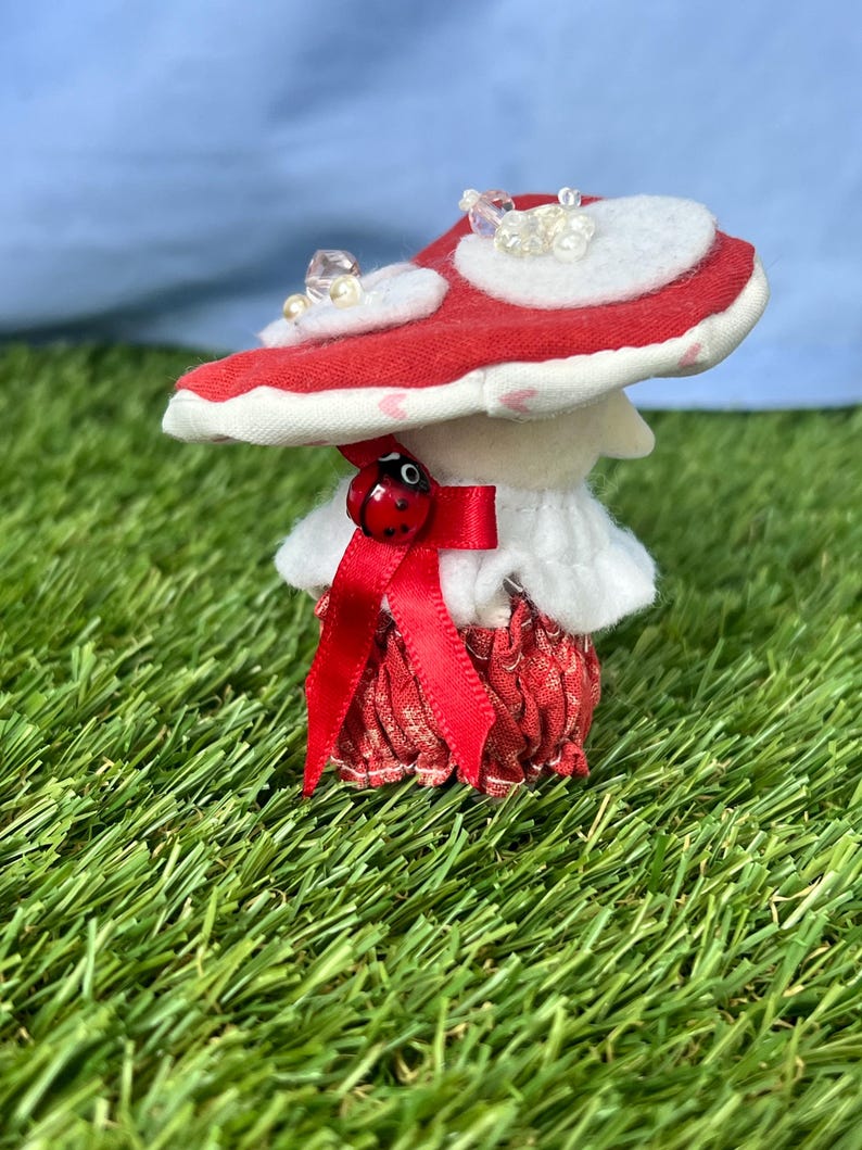 May include: A charming mushroom ornament featuring a red and white cap decorated with clear beads. The mushroom has a white stem, a red skirt, and a red bow with a ladybird detail. It is set against a backdrop of green grass and a blue sky.