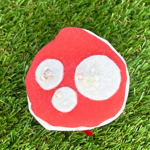 May include: A red mushroom-shaped fabric ornament with three white circular spots. The spots are embellished with small, clear beads. The ornament has a white border and a small red ribbon.