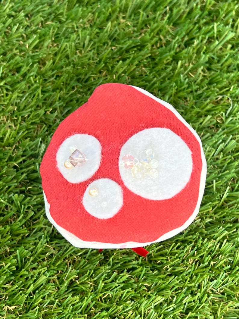May include: A red mushroom-shaped fabric ornament with three white circular spots. The spots are embellished with small, clear beads. The ornament has a white border and a small red ribbon.