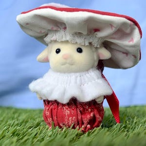 May include: A miniature lamb figure dressed in a red and white mushroom outfit. The hat has a red border and a pattern of small pink hearts. The lamb is set on artificial green grass. The outfit includes a frilly white collar.