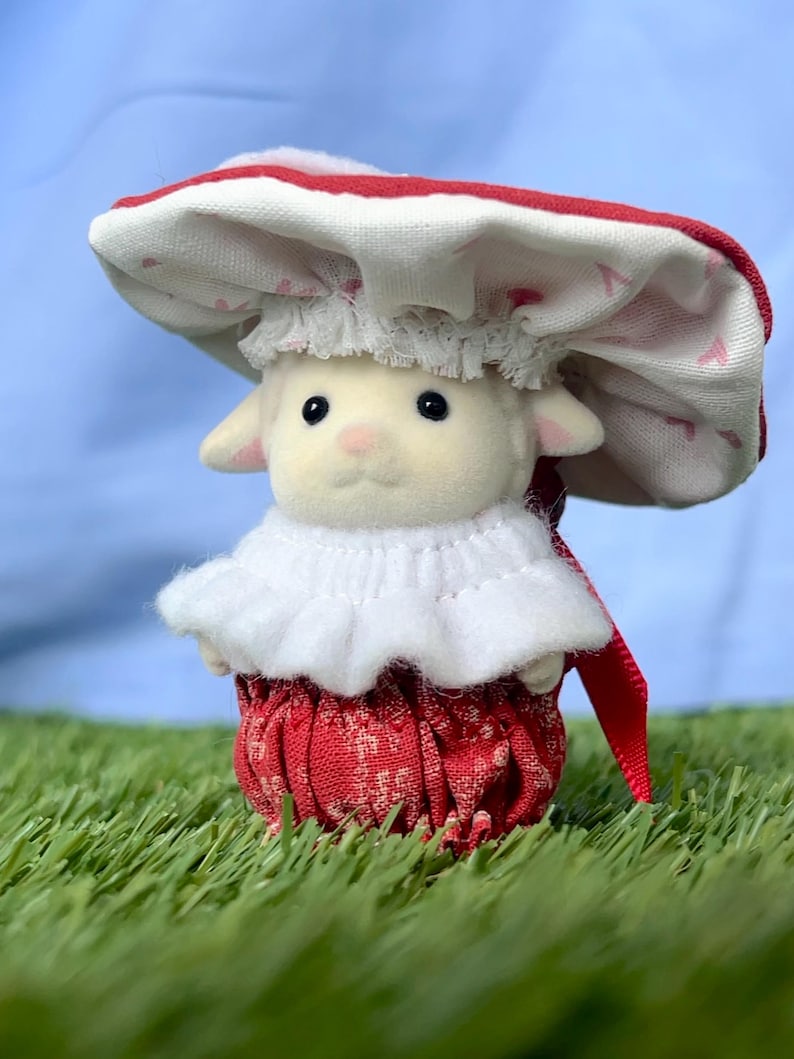 May include: A miniature lamb figure dressed in a red and white mushroom outfit. The hat has a red border and a pattern of small pink hearts. The lamb is set on artificial green grass. The outfit includes a frilly white collar.