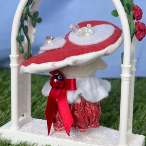 May include: A charming mushroom figure with a red and white cap, embellished with faux pearls, is positioned beneath a white archway decorated with roses. The mushroom wears a red bow with a ladybug detail, a ruffled white collar, and a red patterned skirt.