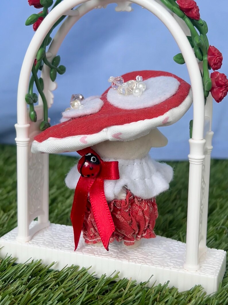 May include: A charming mushroom figure with a red and white cap, embellished with faux pearls, is positioned beneath a white archway decorated with roses. The mushroom wears a red bow with a ladybug detail, a ruffled white collar, and a red patterned skirt.