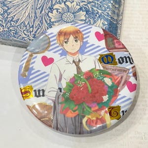 Può includere: Un bottone rotondo con un'illustrazione in stile anime di un personaggio che tiene in mano un mazzo di fiori rossi. Il bottone ha uno sfondo azzurro chiaro con cuori e dettagli dorati. Il testo "The Cat News" è visibile.