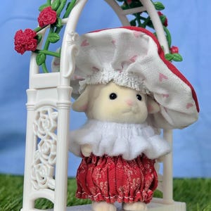 May include: A toy lamb in a red and white patterned dress and bonnet stands within a white decorative arch adorned with red rose detailing. The lamb has a white ruffled collar and is on a white platform.