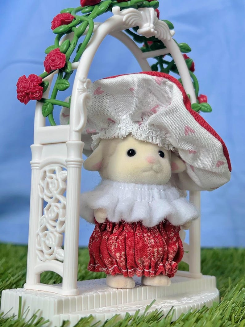 May include: A toy lamb in a red and white patterned dress and bonnet stands within a white decorative arch adorned with red rose detailing. The lamb has a white ruffled collar and is on a white platform.