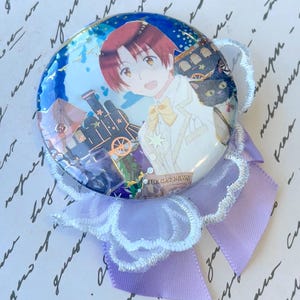 Può includere: Un bottone rotondo in stile anime con un personaggio dai capelli rossi, un gatto nero e un treno. Ha un bordo in pizzo bianco e un nastro lavanda. Il testo "THE CAT NEWS" è visibile.