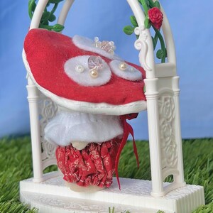 May include: A miniature wedding cake in red and white, decorated with pearls and crystals, sits beneath a white arch. The arch is adorned with red roses and green leaves, creating a romantic, decorative scene.