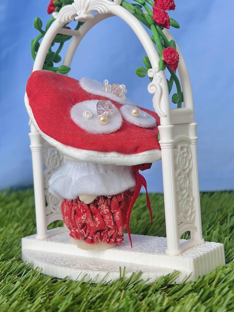 May include: A miniature wedding cake in red and white, decorated with pearls and crystals, sits beneath a white arch. The arch is adorned with red roses and green leaves, creating a romantic, decorative scene.