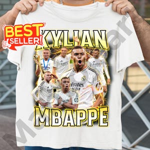 May include: White t-shirt featuring a graphic of Kylian Mbapp&eacute; with the text "KYLIAN MBAPPE". The design includes multiple images of the soccer player in action, with a "Best Seller" sticker in the upper left corner.