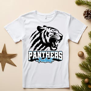 Puede incluir: Camiseta blanca con un diseño de tigre en blanco y negro y el texto "PANTHERS Football" en azul y blanco. El tigre está rugiendo. La camiseta tiene cuello redondo.