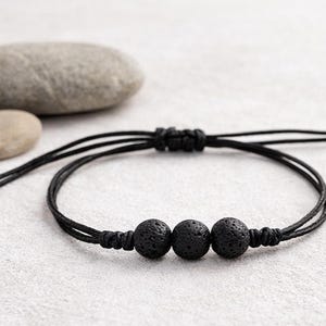 May include: A black cord bracelet featuring three spherical, porous black lava stone beads centered on the band. The bracelet includes an adjustable sliding knot closure with two thin black cords extending from the sides, finished with small knotted ends.
