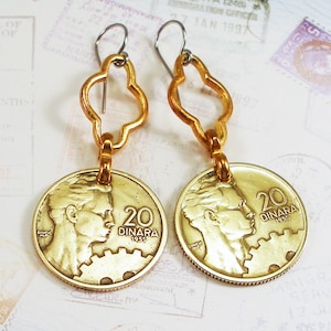 May include: Gold dangle earrings featuring a coin charm with a profile portrait and the text "20 DINARA 1955". The coin is attached to a gold clover-shaped charm.