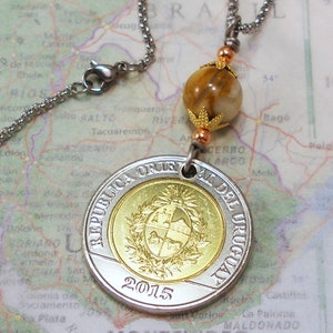 K&ouml;nnte beinhalten: Eine silberne Kettenkette mit einem goldenen und silbernen M&uuml;nzenanh&auml;nger mit dem Text "REPUBLICA ORIENTAL DEL URUGUAY" und dem Jahr "2015". Die M&uuml;nze ist an einer braunen und goldenen Perle mit sternf&ouml;rmigem Design befestigt.