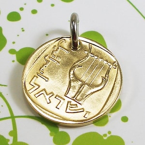 May include: A gold-colored pendant featuring a lyre, a stringed musical instrument, with the Hebrew word "Shabbat" inscribed around the edge.