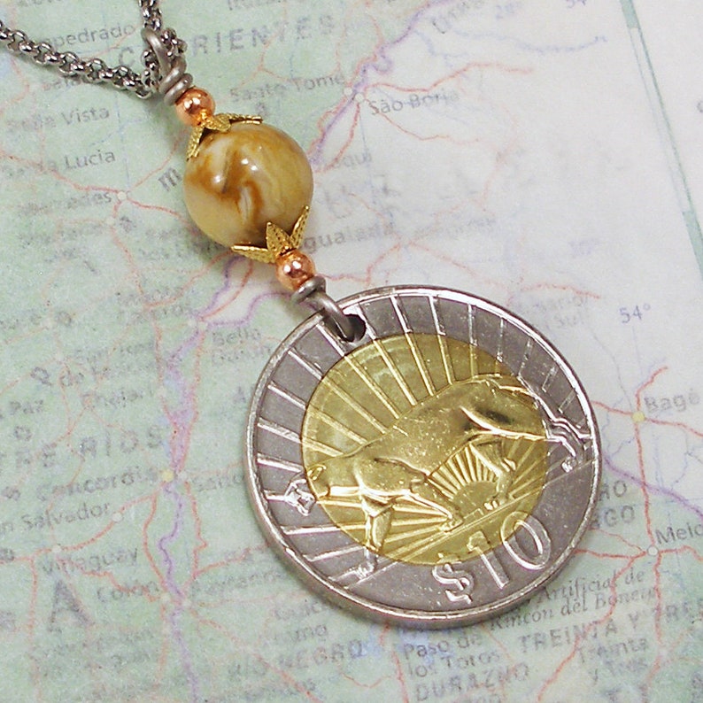 K&ouml;nnte beinhalten: Eine Halskette mit einem Anh&auml;nger aus einer goldenen und silbernen M&uuml;nze, verziert mit einer braunen und wei&szlig;en Marmorperle. Die M&uuml;nze zeigt ein stilisiertes Bild eines Stiers und den Text "$10".