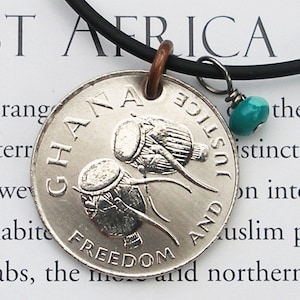 May include: A silver coin necklace with a black cord. The coin features a Ghanaian design with two drums and the words "Ghana" and "Justice and Freedom". A turquoise bead hangs from the cord.