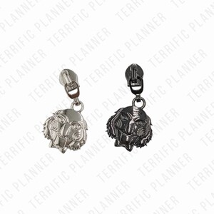 May include: Two zipper pulls with a metal finish. One is silver and the other is black. Both have a detailed design of a face with a wide mouth and large eyes.