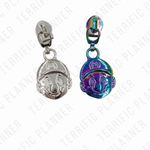 May include: Two zipper pulls featuring a cartoon character with a red cap and mustache. One zipper pull is silver, and the other is iridescent.