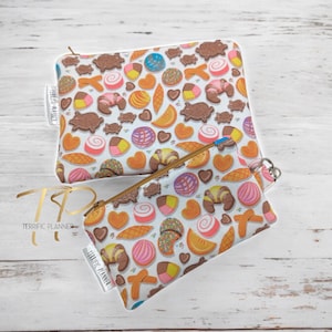 May include: Two white zippered pouches with a colorful pattern of various Mexican pastries and sweets. The pouches are made of fabric and have a white background with a brown zipper. The text "Terrific Planner" is printed on the pouches.