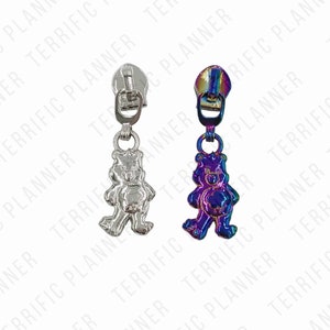May include: Two zipper pulls with bear charms. One is silver-toned, the other has a rainbow iridescent finish. Both have a metal zipper pull attached to the top of the bear charm. The background has a repeating pattern of the words "TERRIFIC PLANNER".