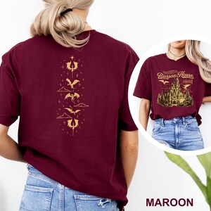 May include: Maroon t-shirt with a gold dragon graphic on the back and a castle design on the front. The back design features a vertical arrangement of stylized dragons and other symbols. The front design includes the text "Riorrson House" and "Aretta".
