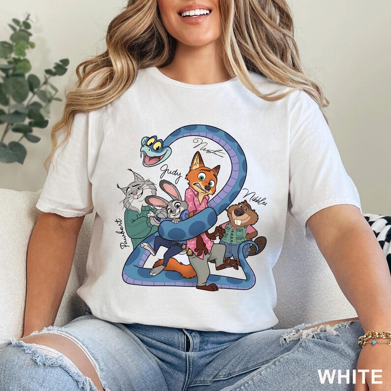Puede incluir: Una camiseta blanca de manga corta con un estampado gr&aacute;fico colorido de personajes de dibujos animados, incluyendo un zorro, un conejo, un lince, un castor y una serpiente azul. Los nombres Pawbert, Judy, Nick y Nibbles aparecen en negro.
