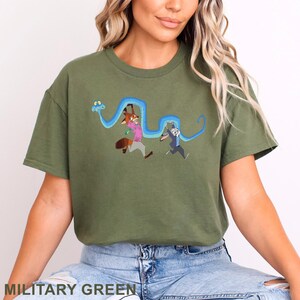 May include: An olive green short-sleeved t-shirt featuring a graphic print of a long blue snake winding around a cartoon fox wearing a pink shirt and a cartoon rabbit wearing a blue suit. The characters appear to be running while holding the snake.
