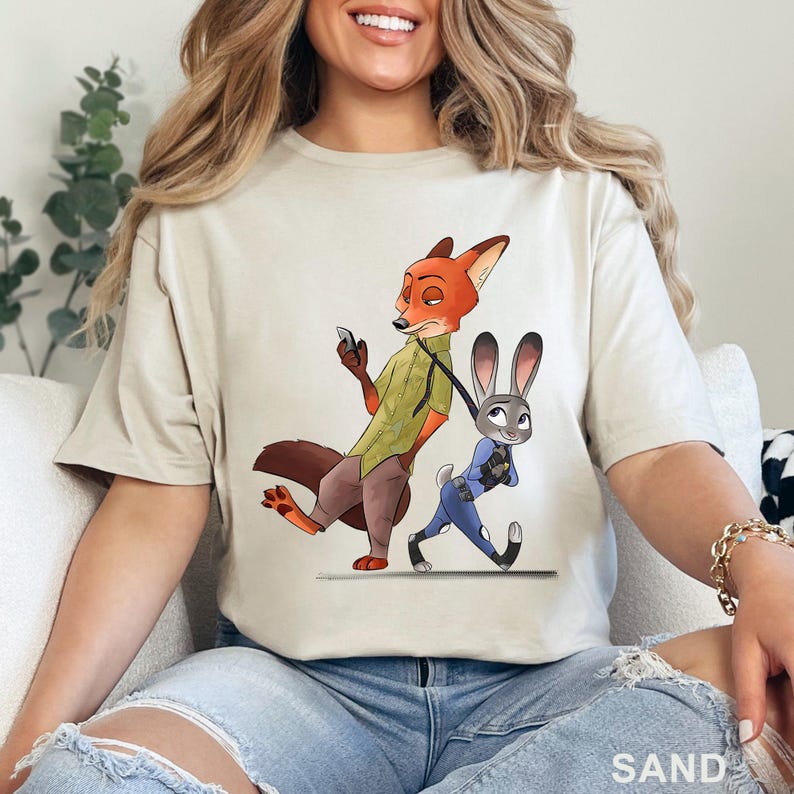 May include: Sand-colored t-shirt featuring a cartoon illustration of Nick Wilde, a fox, and Judy Hopps, a rabbit, from Zootopia. Nick is holding a phone, and Judy is walking. The word "SAND" is printed at the bottom.
