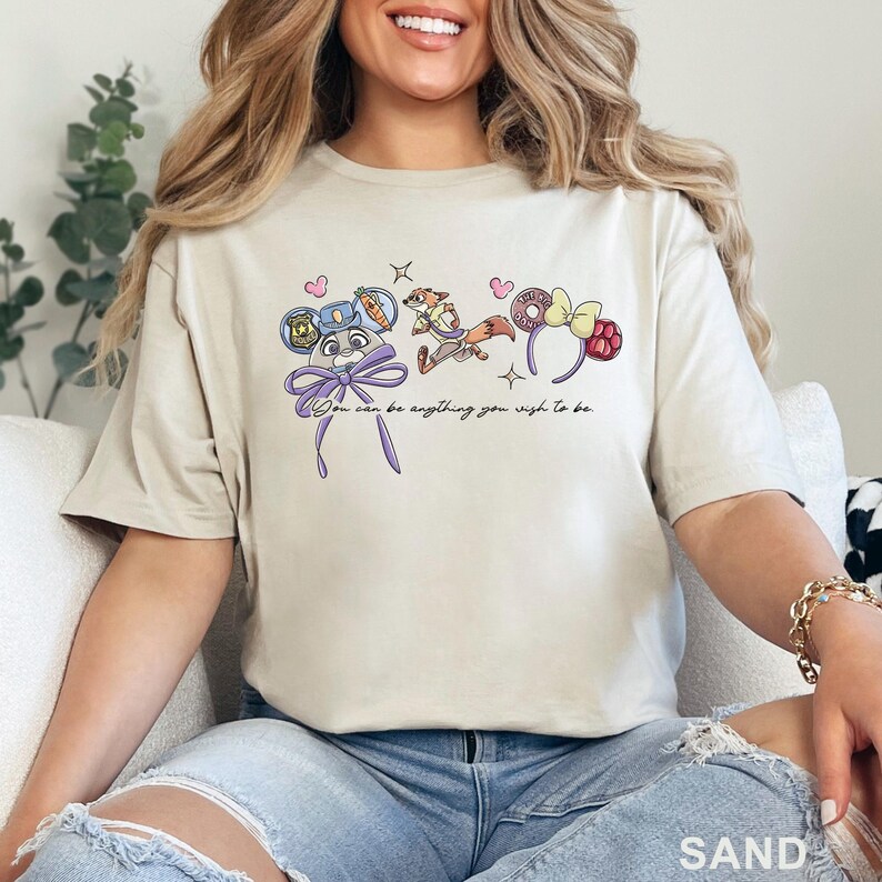 May include: A sand-colored t-shirt featuring a graphic print of a cartoon rabbit, a fox, and two pairs of mouse ears. The text "You can be anything you wish to be" is printed in black cursive script below the illustrations, accented by small pink hearts.