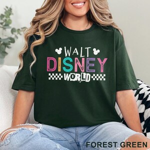 Pu&ograve; includere: Una t-shirt a maniche corte verde bosco con la scritta WALT DISNEY WORLD in lettere colorate. Il design include due piccole sagome bianche di teste di Topolino e un bordo a scacchi bianco sotto la parola WORLD.