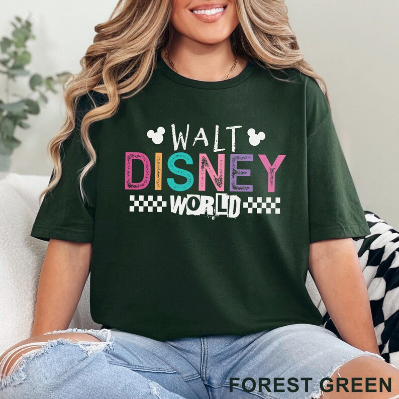 Pu&ograve; includere: Una t-shirt a maniche corte verde bosco con la scritta WALT DISNEY WORLD in lettere colorate. Il design include due piccole sagome bianche di teste di Topolino e un bordo a scacchi bianco sotto la parola WORLD.