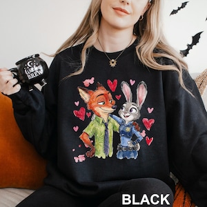 May include: Black sweatshirt with a colourful illustration of Nick Wilde and Judy Hopps from Zootopia, surrounded by red hearts. The word "BLACK" is printed at the bottom. A black mug with the text "Witches Brew" is held in the image.