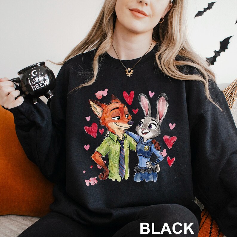 May include: Black sweatshirt with a colourful illustration of Nick Wilde and Judy Hopps from Zootopia, surrounded by red hearts. The word "BLACK" is printed at the bottom. A black mug with the text "Witches Brew" is held in the image.