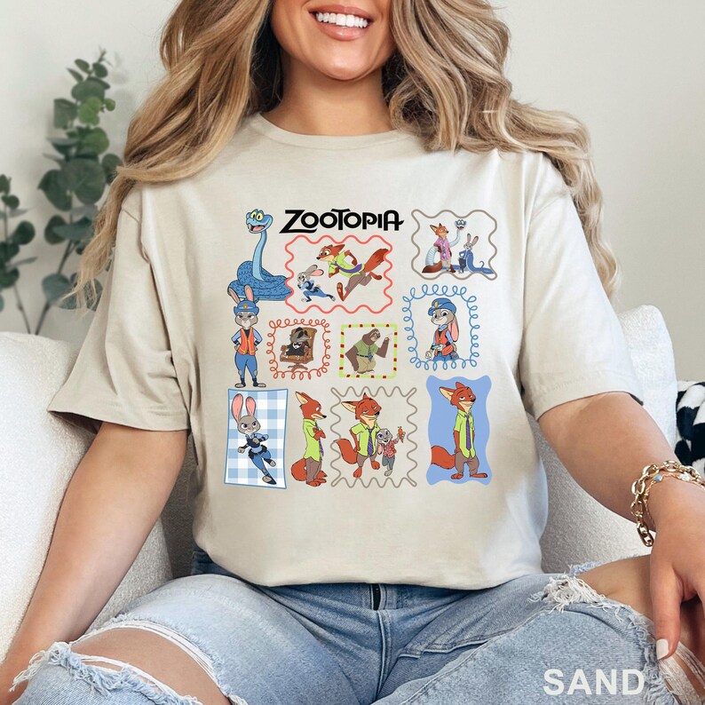 K&ouml;nnte beinhalten: Ein sandfarbenes T-Shirt mit einem Grafikdruck verschiedener Zootopia-Figuren, darunter Judy Hopps und Nick Wilde, in mehreren gerahmten Szenen. Das Wort Zootopia ist in Schwarz oben in der Mitte der farbenfrohen Charakter-Collage gedruckt.