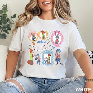 Pode incluir: Camiseta branca com ilustra&ccedil;&otilde;es de desenhos animados coloridos de personagens do filme da Disney Zootopia. O design inclui o texto "Welcome to Zootopia" e imagens emolduradas dos personagens. A camisa est&aacute; rotulada "WHITE" na parte inferior.